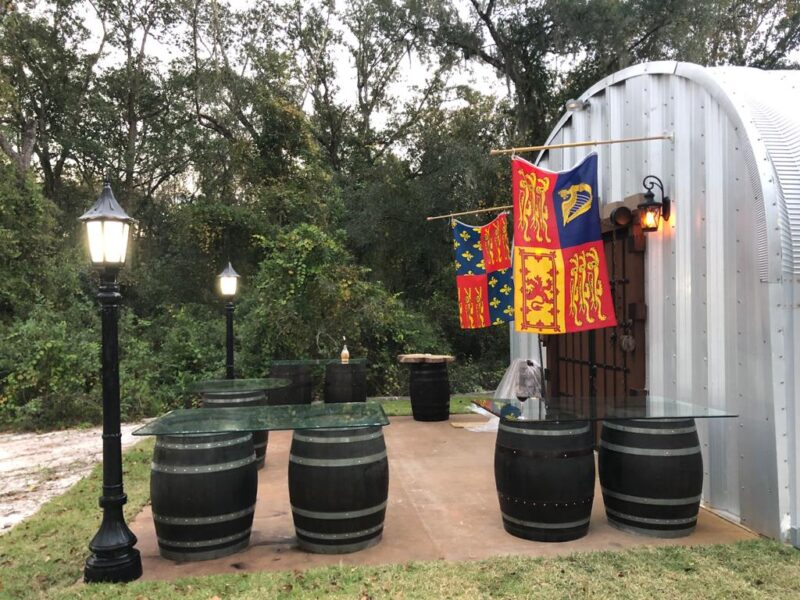 S-model Quonset hut with metal endwall, brown medieval entrance doors and flags, black barrel tables with glass tops in front