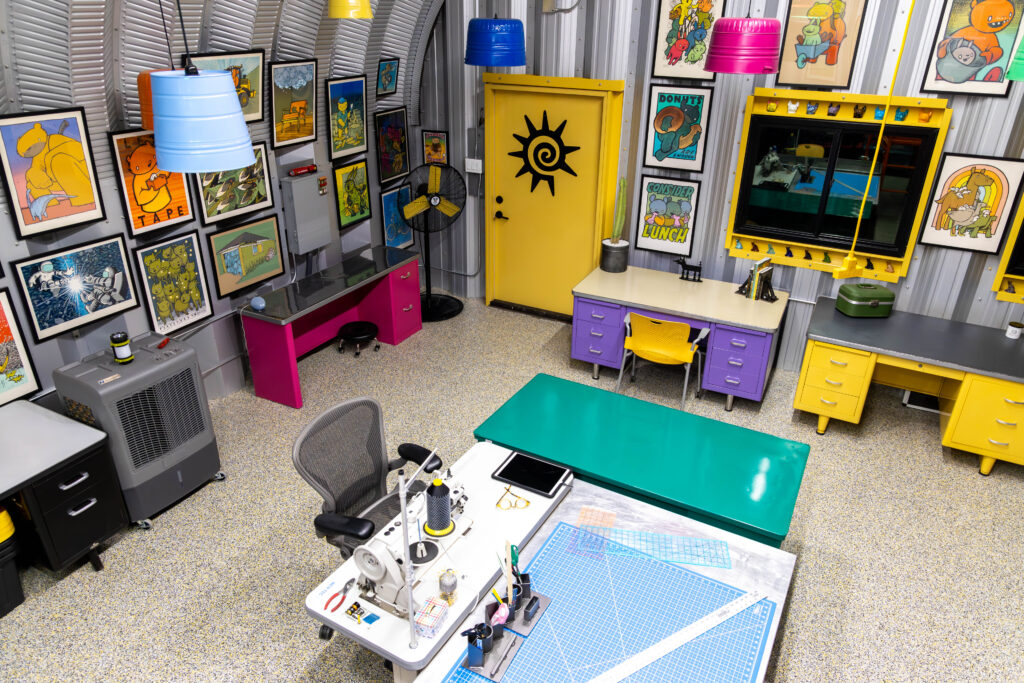 Inside of an S-Model Quonset hut with paintings all over the walls, a teal table, yellow door with a black symbol, and three desks of different colors.