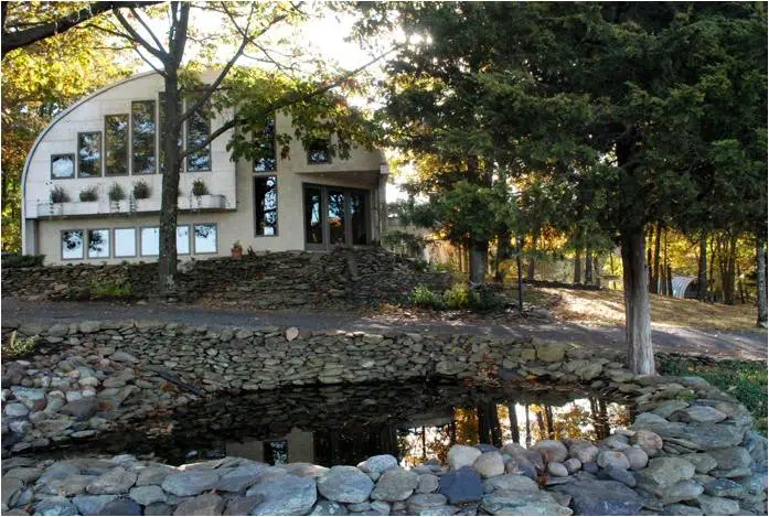 Eco-friendly S-model Quonset home in the woods with a custom tan endwall, large windows, and a small pond out front.