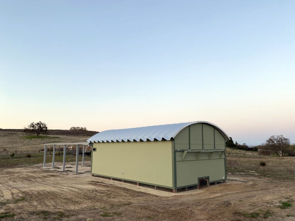 Small, yellow and green trimmed observatory building with steel roof on raised foundation.