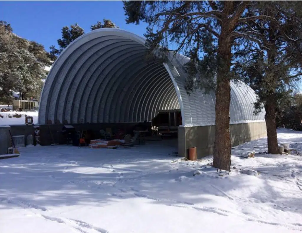 Open ended Quonset structure set on raised concrete foundation, sheltering machinery and equipment, snowy environment.