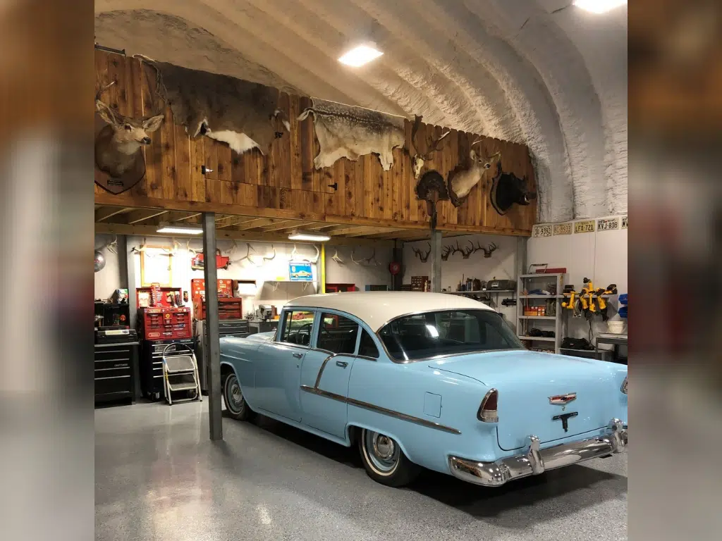 Inside of Quonset workshop: blue vintage car, wooden loft with hanging animal mounts and skins, antlers, tools and shelving lining walls.