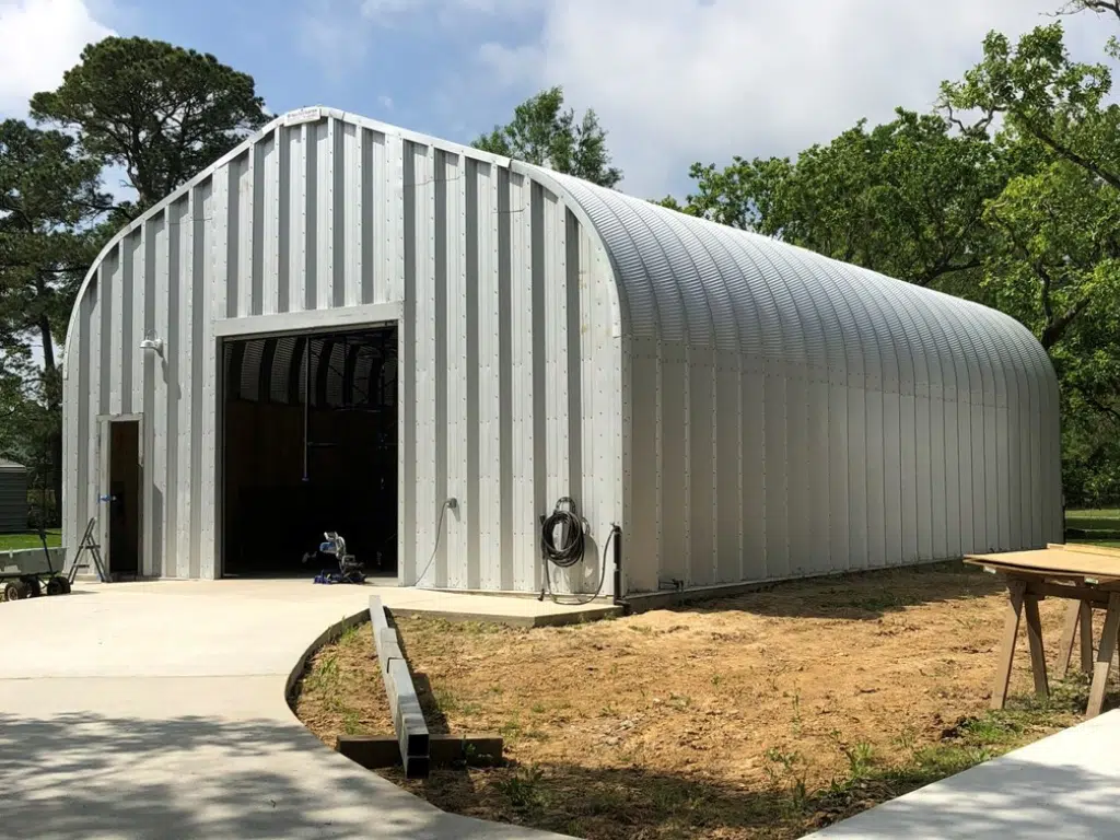 Driveway leading to A-model Quonset hut with steel endwalls, rolling door, front entrance door, hanging water hose.