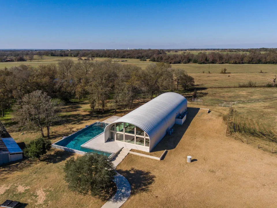 Aerial view of S-Model house with recessed white end wall and large windows, several side windows, and large L-shaped pool